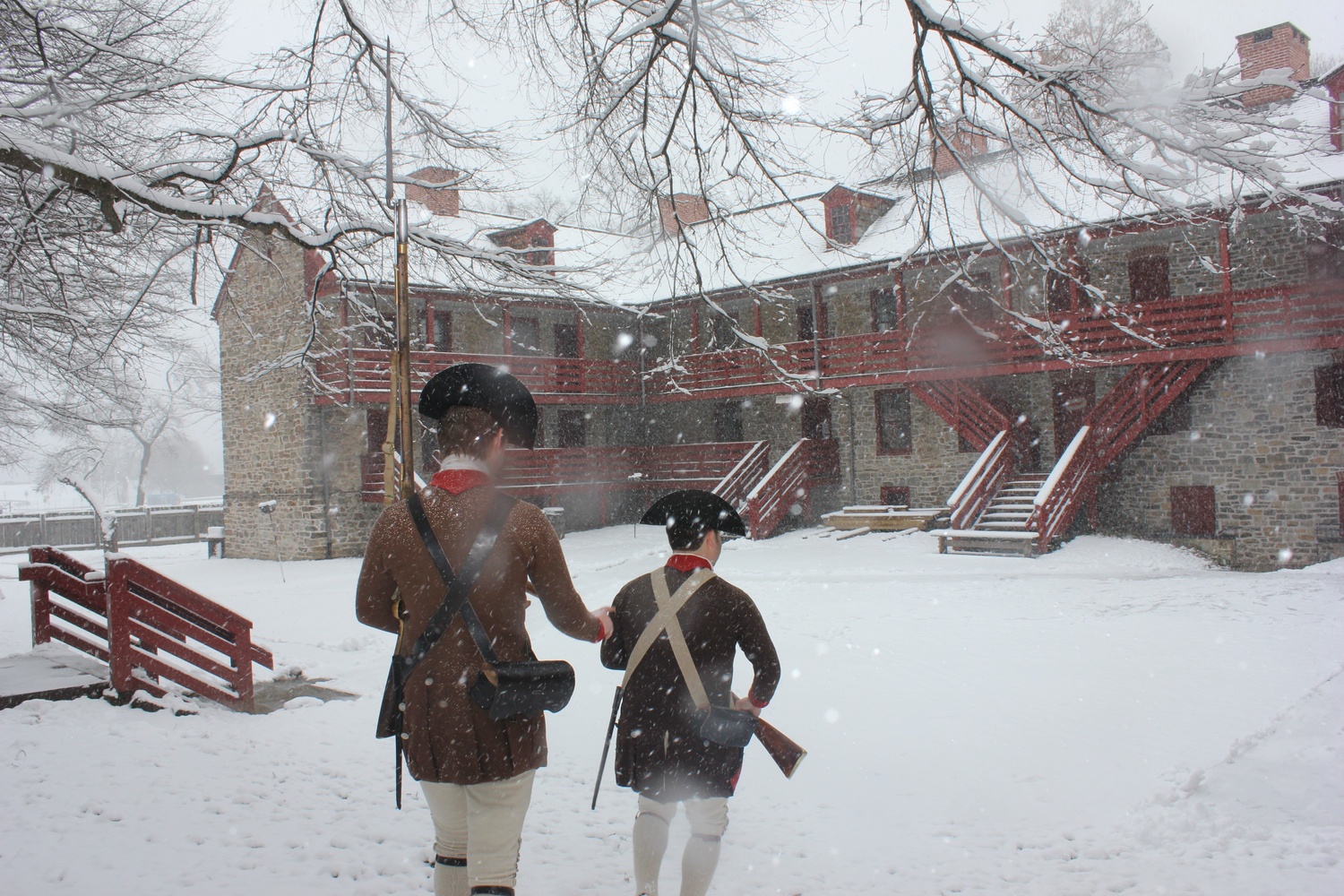 Two people in period clothing stand outdoors in winter at a historic site during a reenactment event.