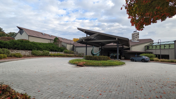 Modern building entrance with covered walkway, landscaping, and paved driveway under a cloudy sky.