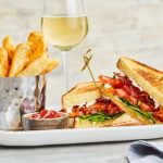 Crispy fries, toasted sandwich with bacon and greens, and white wine on a platter.