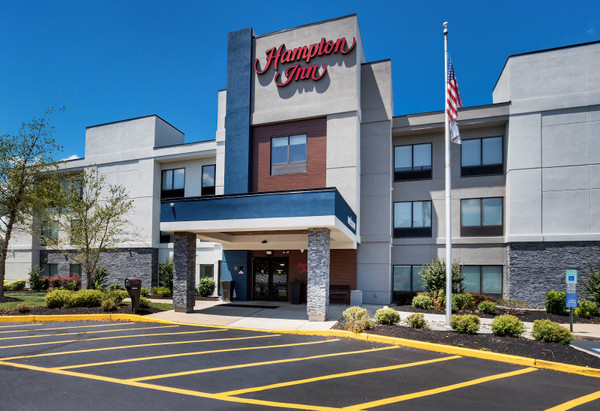 Modern Hampton Inn hotel exterior with a large parking lot and clear blue sky.