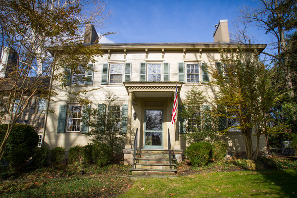 Historic two-story colonial house with green shutters, set on a grassy lawn under blue sky.