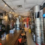 Brewery interior with large stainless steel fermentation tanks and a wooden bar counter.