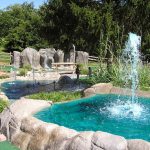 Outdoor splash park with a large fountain, rock formations, and lush greenery.