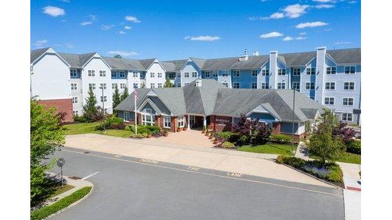 Large apartment complex under a bright blue sky with surrounding greenery and paved road.