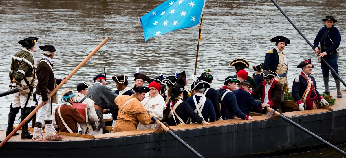 Sailors in historical dress aboard a dark boat on the water under a blue flag.