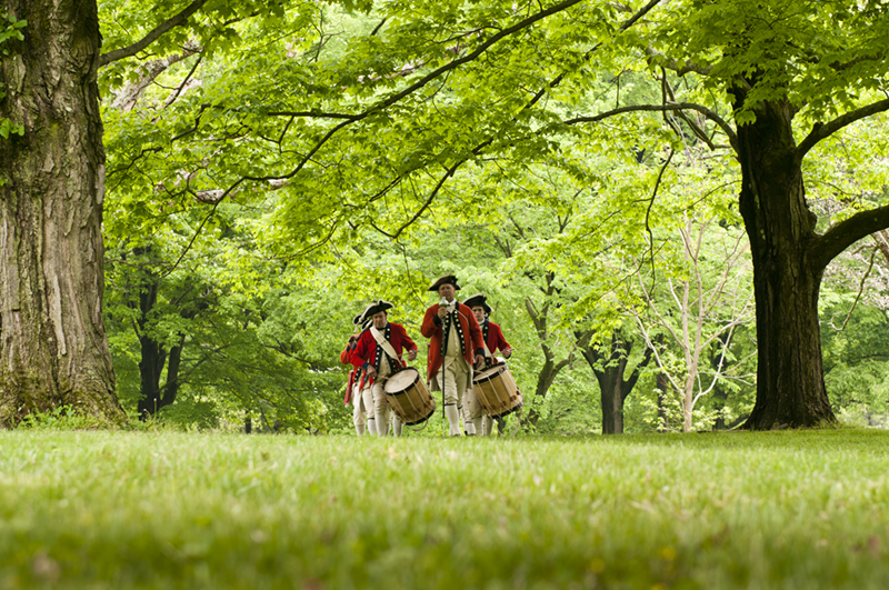 Colonial band members march through a lush, green park under bright daylight.