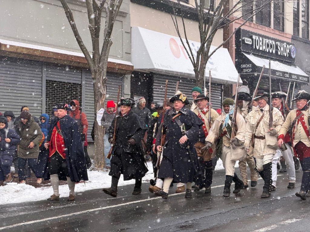 Revolutionary War reenactors march through downtown Trenton during a snowy Patriots Week parade as spectators watch from the sidewalks.