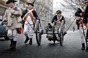 Reenactors in 18th-century Revolutionary War uniforms march down a street, pulling a small wooden cannon on wheels during a historical reenactment.