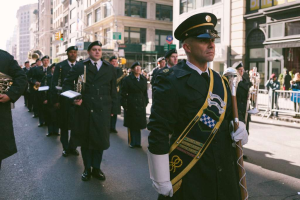 Members of a military marching band participate in a downtown parade, wearing formal uniforms and carrying instruments as they march along a city street lined with spectators.